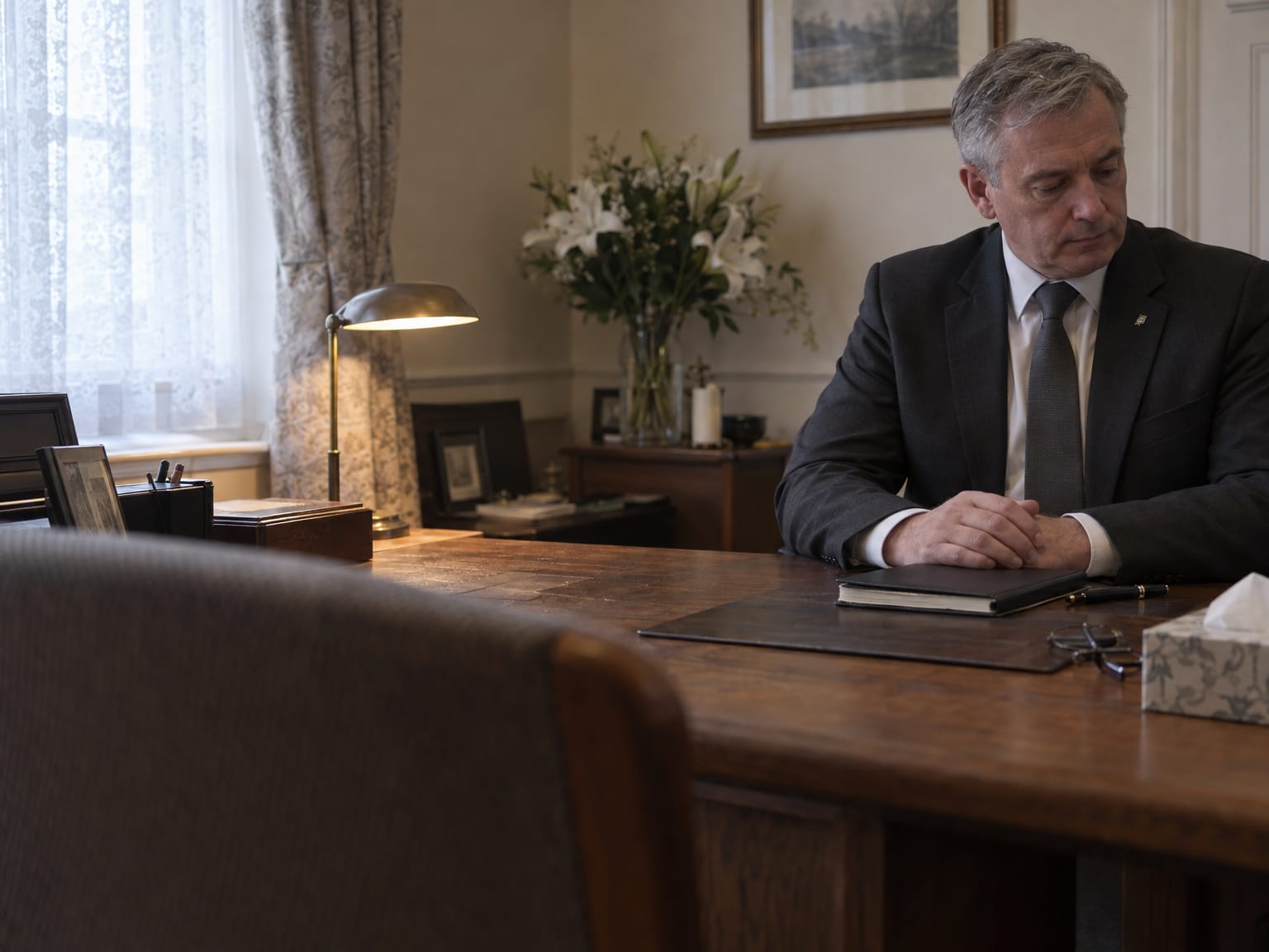 A funeral director in a charcoal suit sitting at an oak desk in a quiet office, hands resting on a closed notebook
