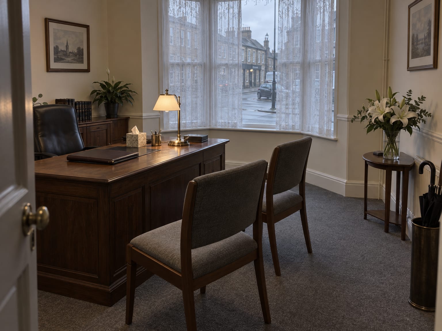 A quiet funeral director’s front office with an oak desk, a lit lamp, two chairs and white flowers by a net-curtained window